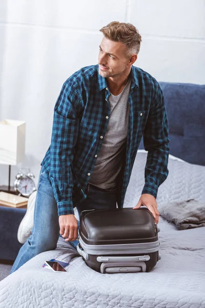 Hombre embalaje bolsa de viaje y mirando hacia otro lado en el dormitorio en casa - foto de stock