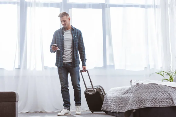 Fröhlicher Mann mit Smartphone und Rolltasche zu Hause — Stockfoto