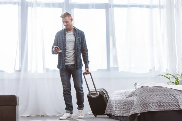 Cheerful man using smartphone and carrying wheeled bag at home — Stock Photo