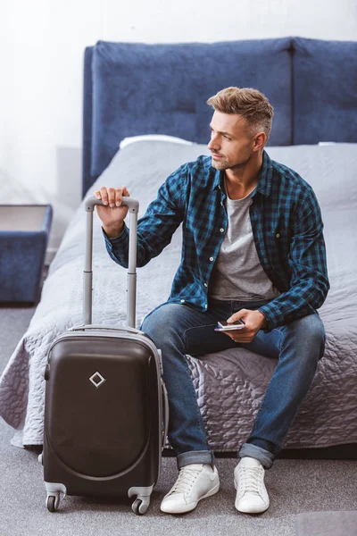 Hombre sentado en la cama con smartphone y billete de avión cerca de la bolsa de ruedas en casa - foto de stock