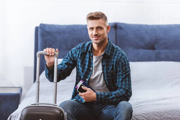 Щасливий чоловік мандрівник сидить на ліжку зі смартфоном і квитком на літак біля колісної сумки вдома — стокове фото