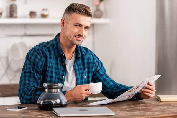 Happy man with coffee cup reading newspaper at table with laptop in kitchen at home — Stock Photo