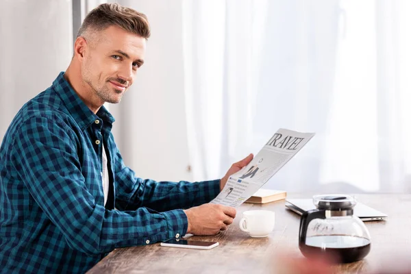 Cheerful man sitting at table and reading travel newspaper in kitchen at home — Stock Photo