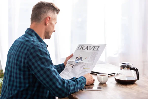 Vista posteriore dell'uomo seduto a tavola e che legge il giornale di viaggio in cucina a casa — Foto stock