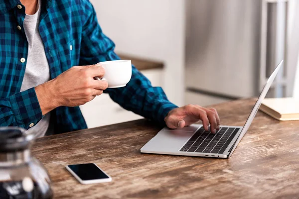 Recortado disparo de hombre sosteniendo la taza de café y el uso de la computadora portátil en casa - foto de stock