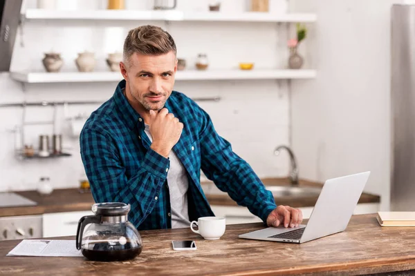 Homem bonito em camisa quadriculada usando laptop e olhando para a câmera — Fotografia de Stock