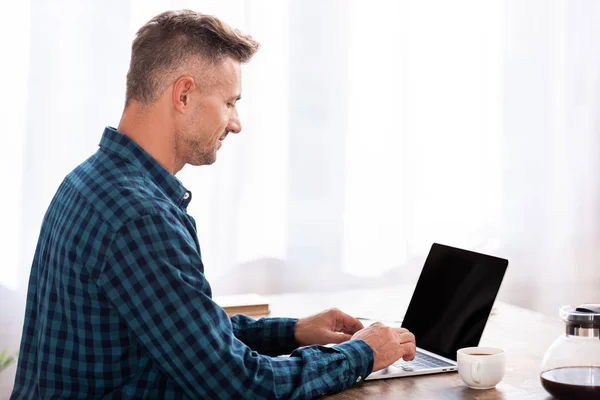 Vista lateral do homem sorridente em camisa quadriculada usando laptop em casa — Fotografia de Stock