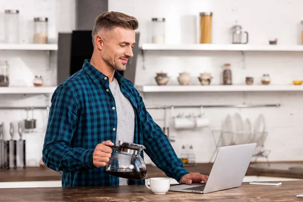 Sonriente hombre sosteniendo cafetera y usando el ordenador portátil en casa - foto de stock