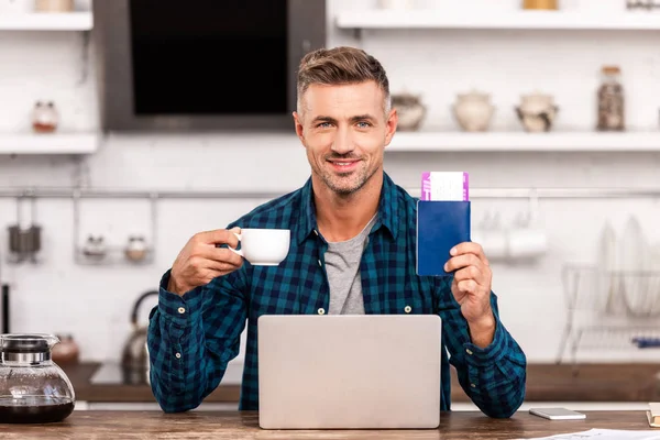 Felice uomo in possesso di una tazza di caffè e passaporto con biglietto durante l'utilizzo del computer portatile a casa — Foto stock