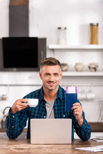 Bonito sorrindo homem segurando xícara de café e passaporte com cartão de embarque ao usar laptop em casa — Fotografia de Stock