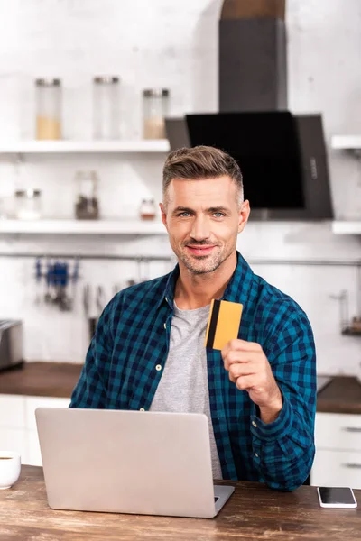 Красивий чоловік тримає кредитну картку і посміхається на камеру, використовуючи ноутбук вдома — стокове фото