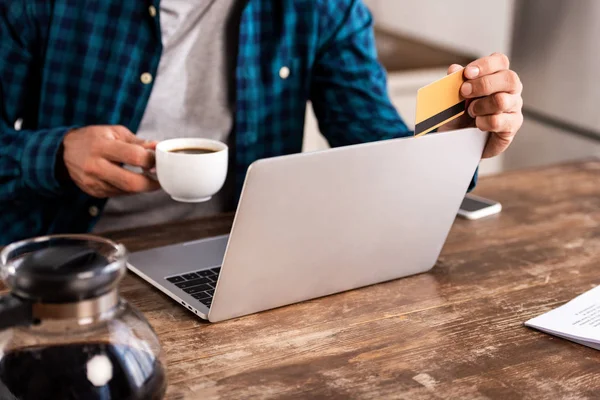 Primer plano vista parcial del hombre sosteniendo la taza de café y tarjeta de crédito mientras usa el ordenador portátil en casa - foto de stock