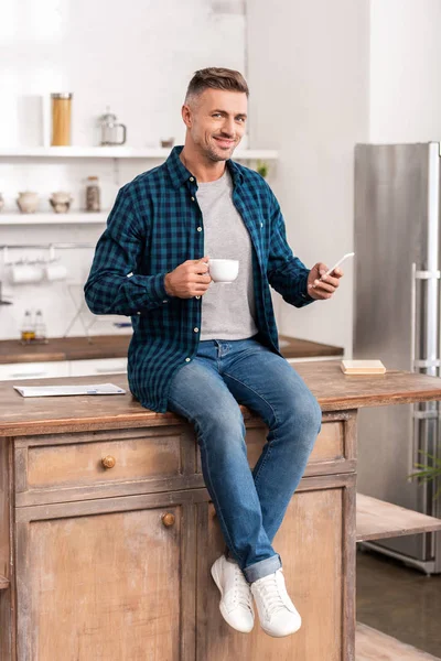 Bell'uomo sorridente che tiene in mano una tazza di caffè e usa lo smartphone mentre siede sul tavolo della cucina — Foto stock