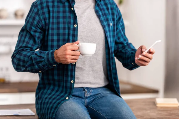 Обрезанный снимок мужчины в клетчатой рубашке с чашкой кофе и с помощью смартфона дома — стоковое фото