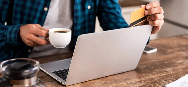 Cropped shot of man holding cup of coffee and credit card while using laptop at home — Stock Photo