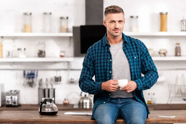 Homem bonito em camisa quadriculada segurando xícara de café e olhando para a câmera na cozinha — Fotografia de Stock