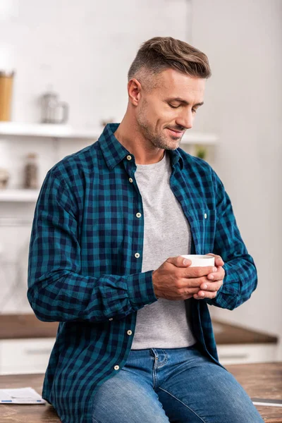 Bonito sorrindo homem em xadrez camisa segurando xícara de café em casa — Fotografia de Stock
