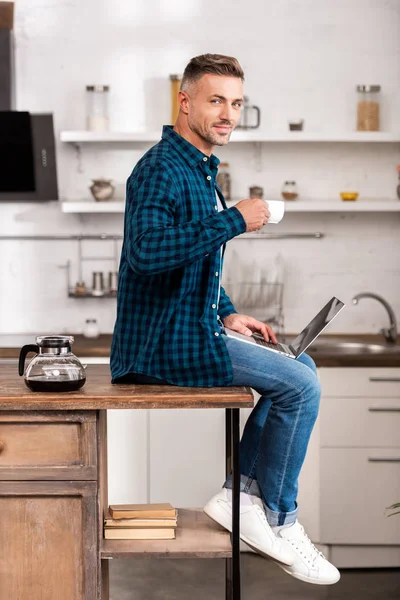 Homem adulto bonito segurando xícara de café e sorrindo para a câmera ao usar laptop em casa — Fotografia de Stock