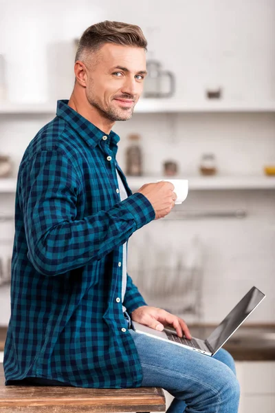 Schöner Mann im karierten Hemd mit einer Tasse Kaffee und einem Lächeln in die Kamera, während er zu Hause den Laptop benutzt — Stockfoto