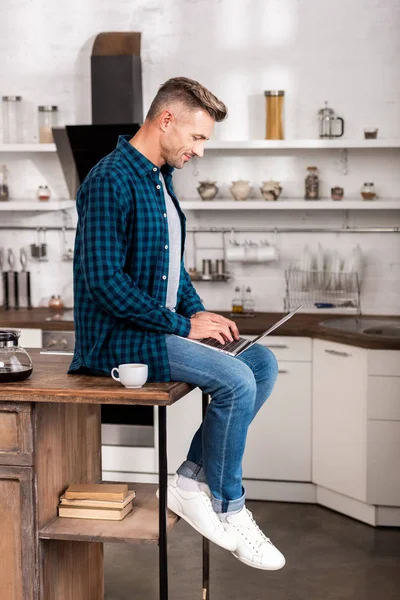 Seitenansicht eines lächelnden Mannes im karierten Hemd, der auf dem Tisch sitzt und zu Hause Laptop benutzt — Stockfoto