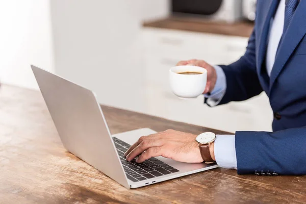Cropped shot of businessman holding cup of coffee and using laptop at home — Stock Photo