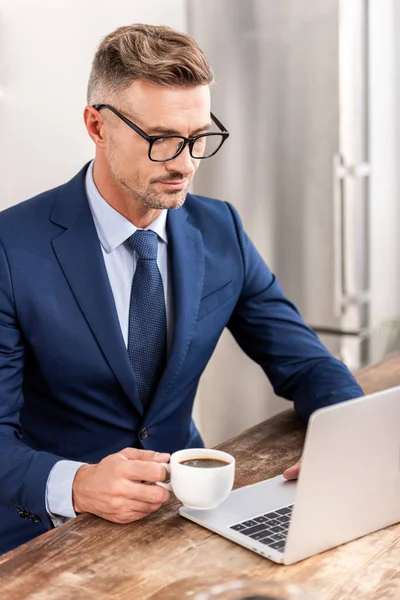 Businessman in eyeglasses drinking coffee and using laptop at home — Stock Photo