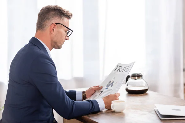 Side view of businessman in suit and eyeglasses reading newspaper at home — Stock Photo