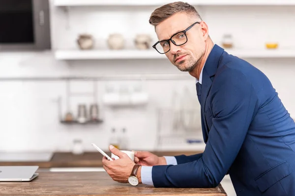 Handsome businessman in eyeglasses using smartphone and looking at camera while drinking coffee in kitchen — Stock Photo