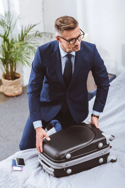 High angle view of businessman in suit and eyeglasses packing suitcase and looking away at home — Stock Photo