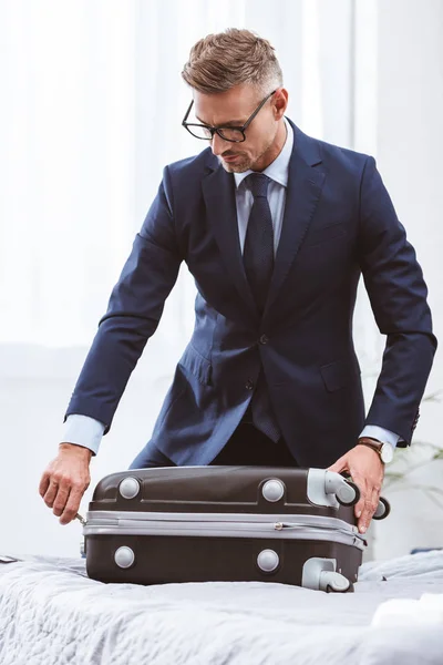 Schöner Geschäftsmann in Anzug und Brille, der Koffer auf dem Bett packt — Stockfoto