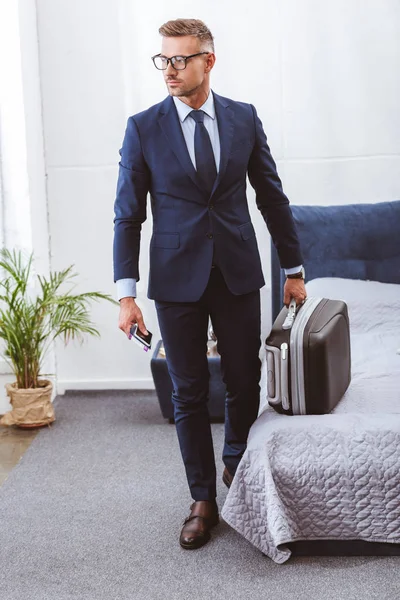 Schöner Geschäftsmann mit Koffer, Smartphone und Reisepass im Schlafzimmer — Stockfoto
