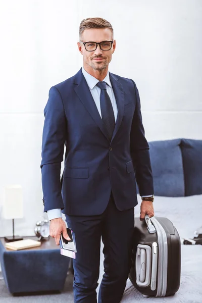 Красивый улыбающийся бизнесмен с чемоданом, паспортом с билетом и смартфоном — стоковое фото