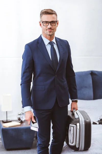 Guapo hombre de negocios sonriente sosteniendo la maleta, pasaporte con billete y teléfono inteligente - foto de stock