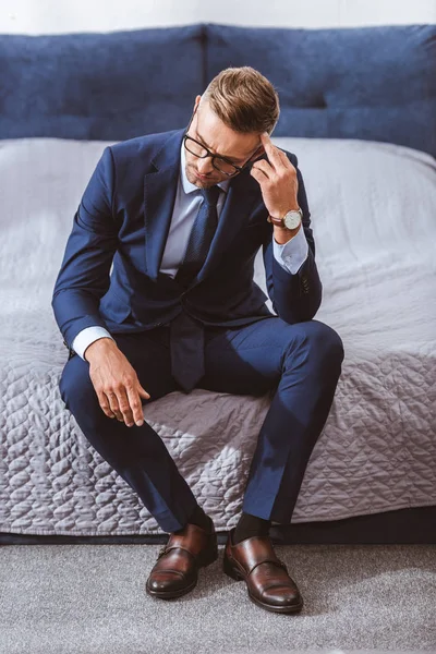 Vue grand angle d'homme d'affaires réfléchi en costume et lunettes assis sur le lit à la maison — Photo de stock