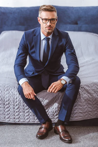 Высокий угол обзора бизнесмена в костюме и очках, сидящего на кровати и смотрящего в камеру — стоковое фото