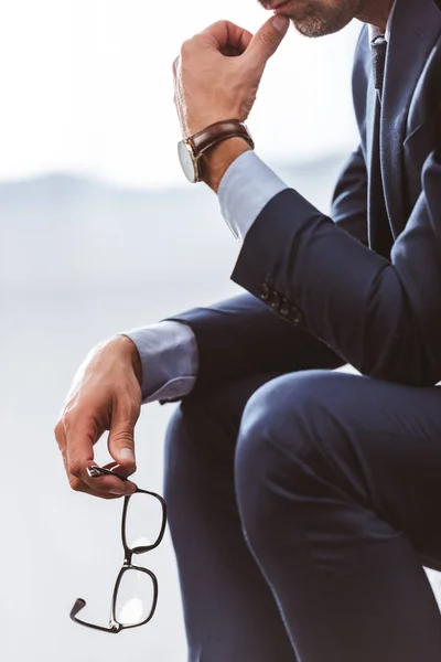 Cropped shot of businessman in suit sitting and holding eyeglasses — Stock Photo