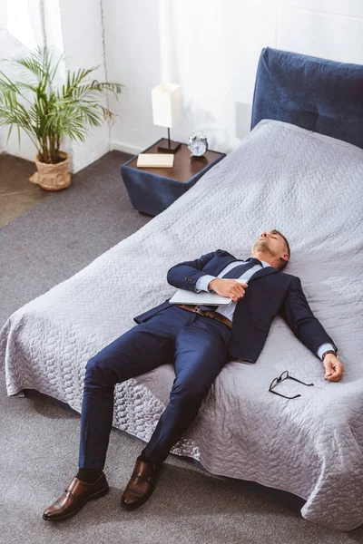 Высокий угол зрения перегруженный бизнесмен держит ноутбук и лежит на кровати — стоковое фото
