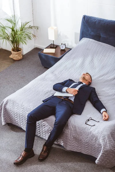 Vista de ángulo alto del hombre de negocios sobrecargado sosteniendo el ordenador portátil y acostado en la cama - foto de stock