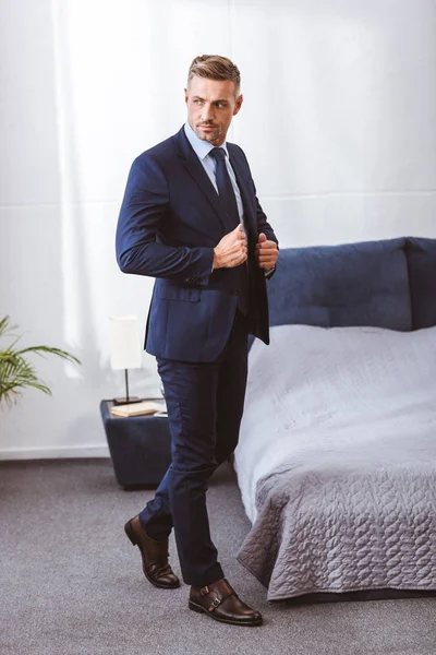 Вид в полный рост на красивого взрослого мужчину в пиджаке и отводящего взгляд в спальне — стоковое фото
