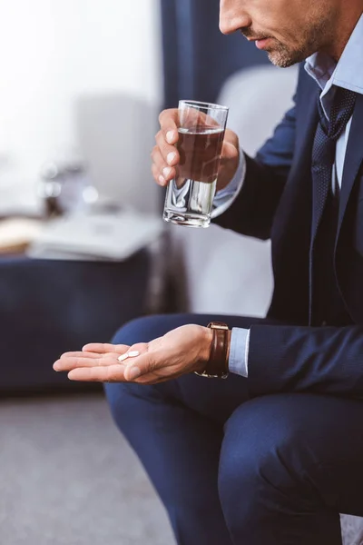Cropped shot of businessman in suit holding glass of water and pills at home — Stock Photo