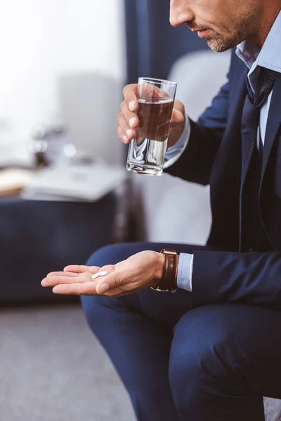 Recortado tiro de hombre de negocios en traje sosteniendo vaso de agua y pastillas en casa - foto de stock