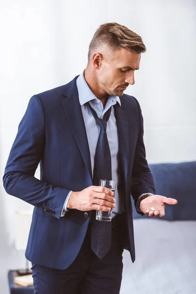 Uomo d'affari in giacca e cravatta con bicchiere d'acqua e pillole a casa — Foto stock