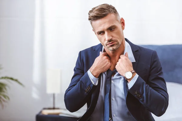 Hombre guapo con corbata y mirando hacia otro lado mientras está sentado en la cama en casa - foto de stock