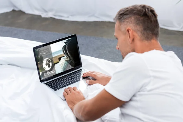 Mann in weißem T-Shirt mit Laptop und Buchungswebseite im Bett — Stockfoto