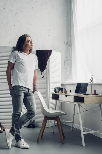 Mujer transexual joven reflexiva de pie cerca del lugar de trabajo y mirando hacia otro lado en casa - foto de stock