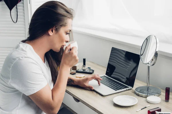 Молодая трансгендерная фрилансер женщина пьет кофе во время работы с ноутбуком на дому — стоковое фото