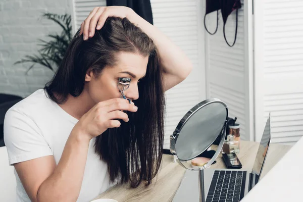 Joven mujer transgénero haciendo maquillaje con rizador de pestañas en casa - foto de stock