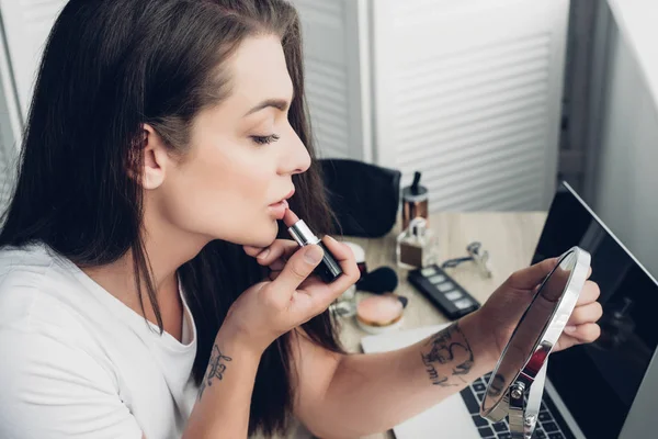 Joven transexual mujer aplicación de lápiz labial mientras mira ronda maquillaje espejo en casa - foto de stock