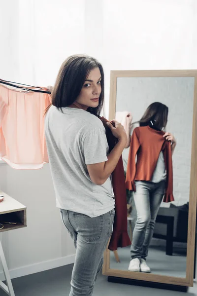 Joven transexual mujer probándose camisas femeninas delante del espejo y mirando a la cámara - foto de stock