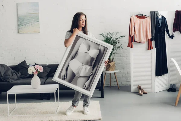 Reflexiva joven transexual sosteniendo foto en blanco y negro con cuerpo femenino en ropa interior en casa - foto de stock